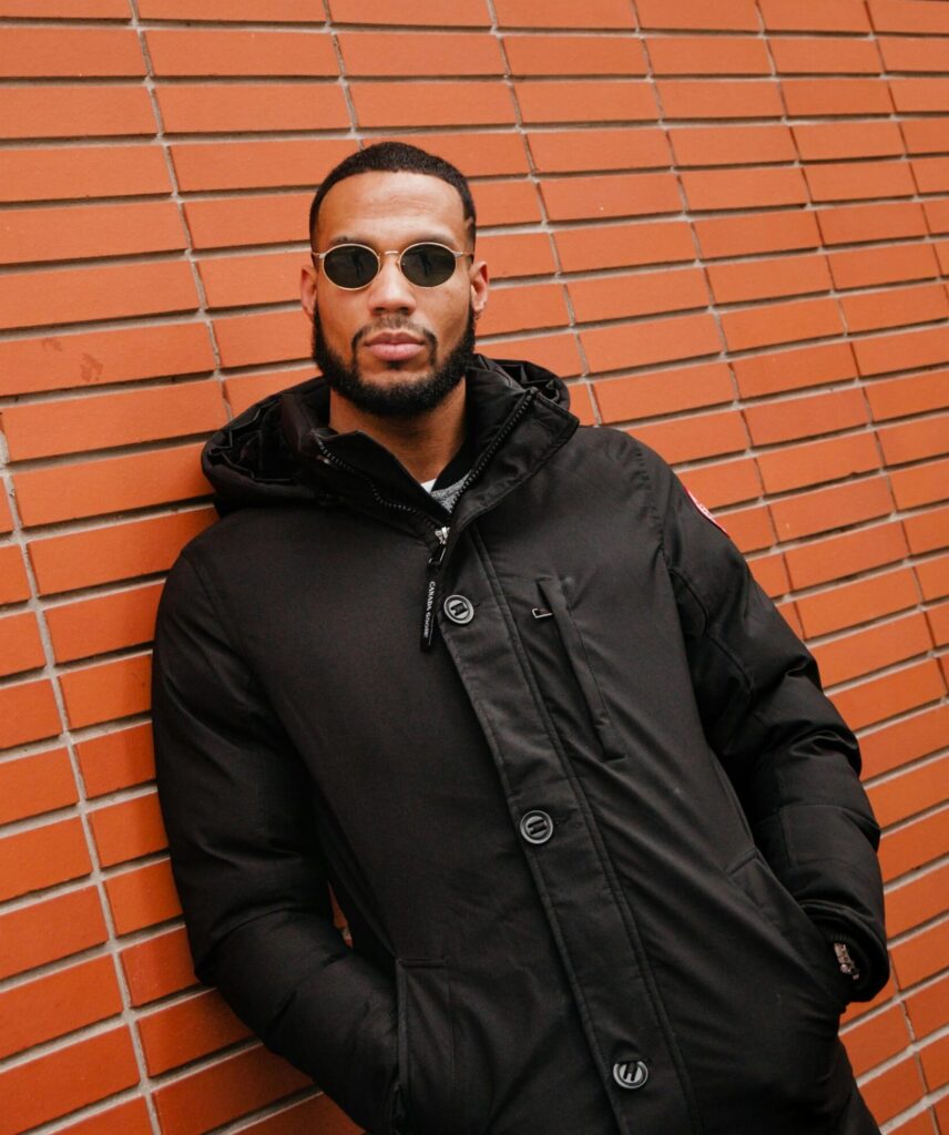 Stylish man wearing sunglasses and black coat leans against a brick wall, embodying urban fashion.