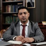 A serious businessman in a suit writing at his desk in a formal office setting.