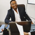 Smiling businessman in suit working with laptop in modern office setting, engaging in discussion.